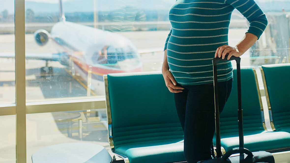 Pregnant woman at an airport, standing beside a window and holding a suitcase, with an airplane visible in the background.