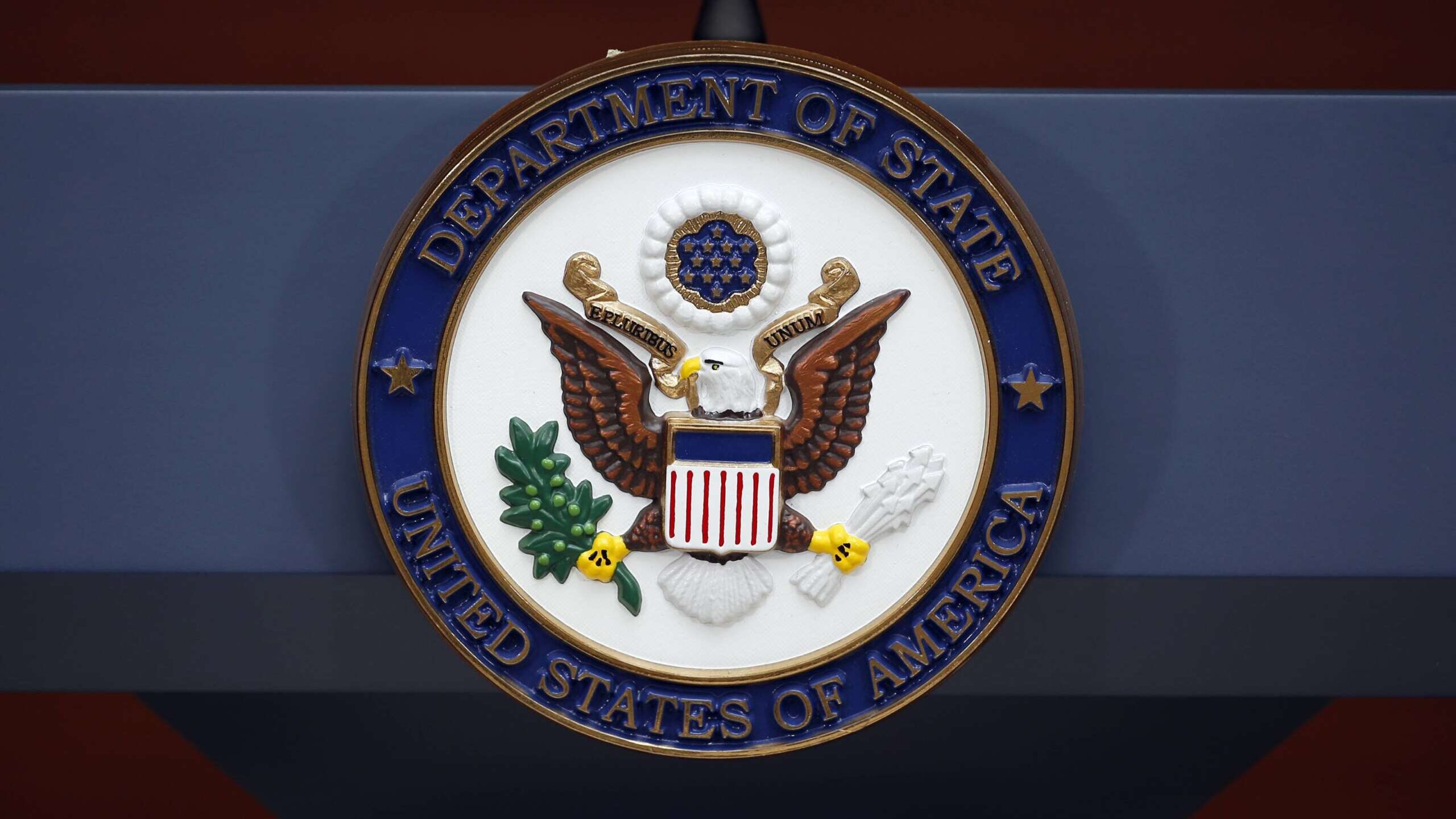 Fotografia em close-up do selo oficial do Departamento de Estado dos EUA, mostrando a águia americana e as inscrições em dourado e azul, montado em um pódio azul escuro.