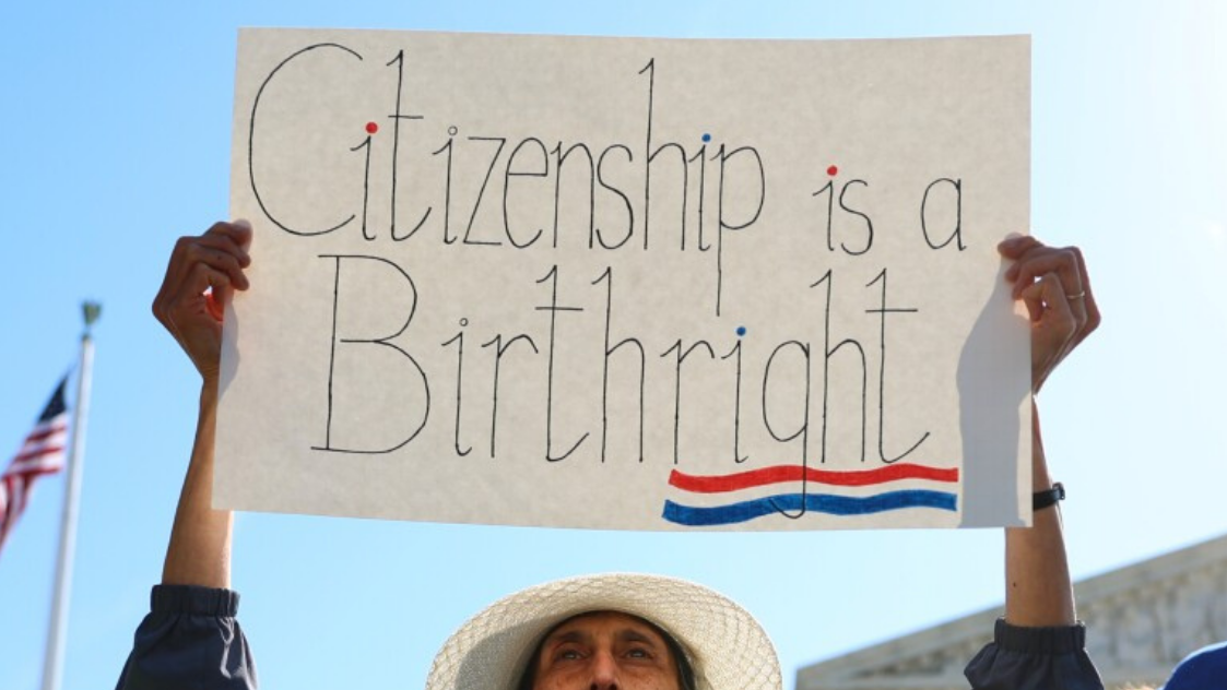 Mulher participa de uma manifestação segurando um cartaz com a frase “Citizenship is a Birthright”, enquanto uma bandeira dos Estados Unidos aparece ao fundo.
