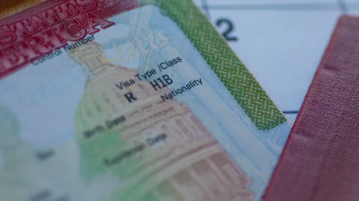 Close-up view of a U.S. visa foil inside a passport, displaying the text "Visa Type / Class R H1B" alongside illustrations of the U.S. Capitol dome