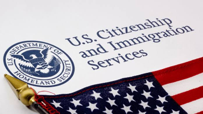 Uma fotografia em plano detalhado de documentos oficiais exibindo o selo circular azul do Departamento de Segurança Interna dos EUA (U.S. Department of Homeland Security) no lado esquerdo. À direita, em fonte escura e em destaque, lê-se o texto: "U.S. Citizenship and Immigration Services". O primeiro plano inferior e o lado direito são ocupados por uma bandeira dos Estados Unidos dobrada, com estrelas e listras visíveis, acompanhada pela ponteira dourada em formato de lança da sua haste.