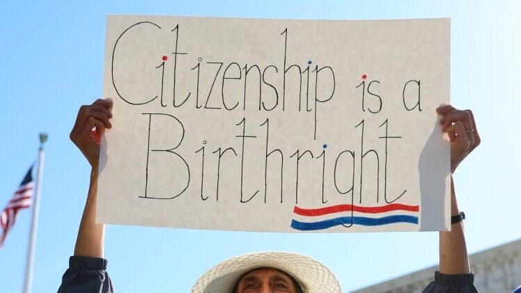 A person holds a sign reading “Citizenship is a Birthright” during a public demonstration, with the U.S. flag and a government building visible in the background.