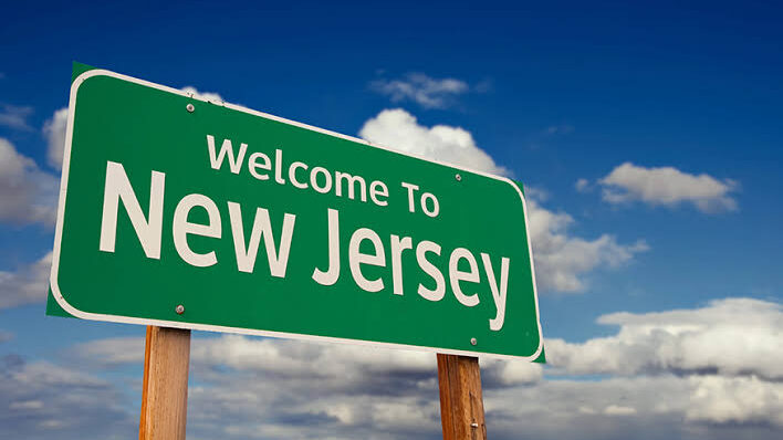 Placa verde de “Welcome to New Jersey” em uma estrada, com o céu azul e nuvens ao fundo.