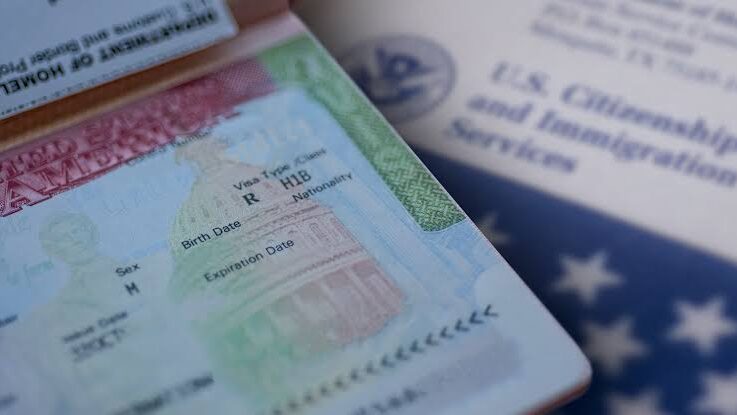 Close-up image of an H-1B U.S. visa in a passport, with U.S. immigration documents and an American flag in the background.