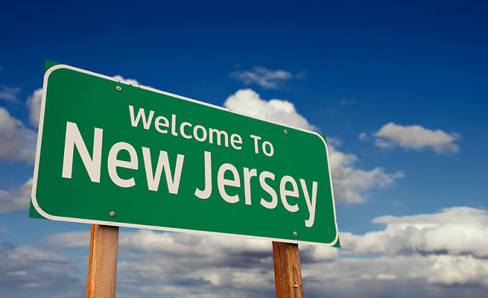 Green highway sign reading “Welcome To New Jersey” against a blue sky with clouds.