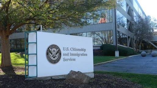 An outdoor view of a U.S. Citizenship and Immigration Services (USCIS) building entrance. In the foreground stands a large white sign with the official U.S. Department of Homeland Security seal on the left and the text "U.S. Citizenship and Immigration Services" in grey lettering to the right. Behind the sign is a multi-story office building with large glass windows reflecting trees and an American flag, surrounded by green lawns and mature trees.