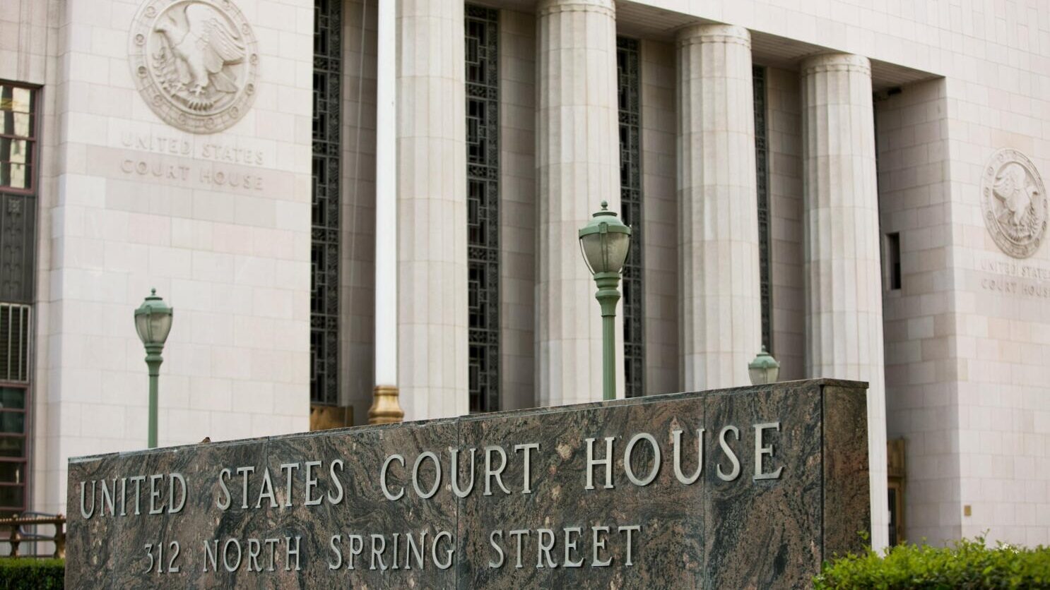A street-level outdoor view of the United States Court House at 312 North Spring Street. In the foreground, a large, dark marbled stone sign reads "UNITED STATES COURT HOUSE" on the top line and "312 NORTH SPRING STREET" below it. Behind the sign is the historic federal building featuring massive stone columns, ornamental green streetlamps, and high vertical windows. Two circular stone bas-reliefs depicting the United States eagle seal are mounted on the facade, with "UNITED STATES COURT HOUSE" carved into the stone below them. Green bushes and a few red flowers are visible at the bottom of the frame