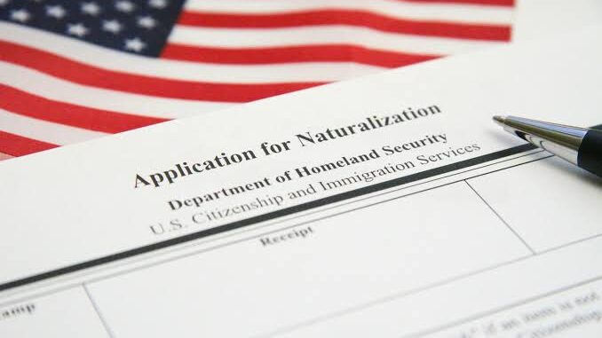 Formulário oficial de solicitação de naturalização dos Estados Unidos (Application for Naturalization) com uma caneta sobre o papel e a bandeira americana ao fundo.
