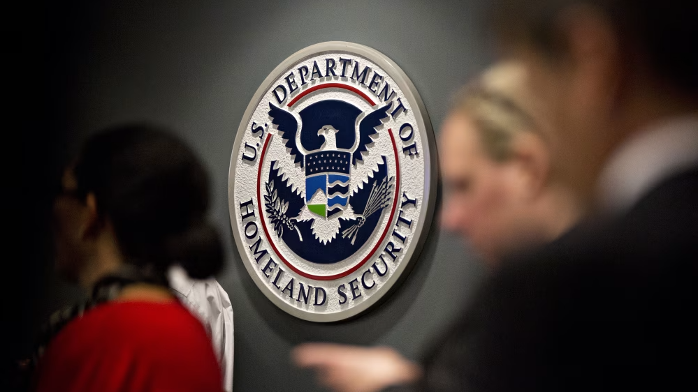 O selo oficial do Departamento de Segurança Interna dos Estados Unidos (U.S. Department of Homeland Security) fixado numa parede cinzenta, com silhuetas desfocadas de pessoas em primeiro plano.