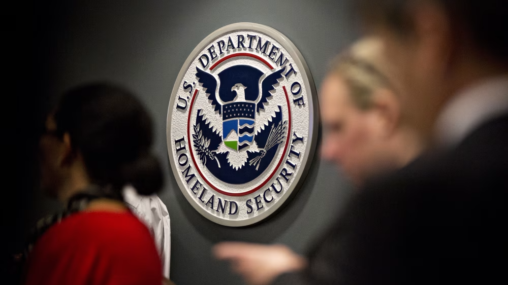 Official seal of the U.S. Department of Homeland Security on a grey wall with blurred figures of people in the foreground.