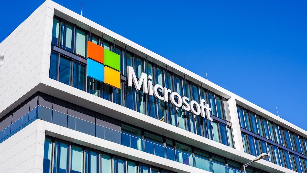 Exterior de um edifício moderno de escritórios da Microsoft, com o logotipo colorido da empresa em primeiro plano. O edifício é feito de vidro e metal, com grandes janelas que refletem o céu azul e a vegetação circundante. O logotipo da Microsoft, com seus quatro quadrados coloridos, está localizado no canto superior esquerdo da fachada do edifício. A foto é tirada de baixo para cima, dando uma sensação de escala e grandeza