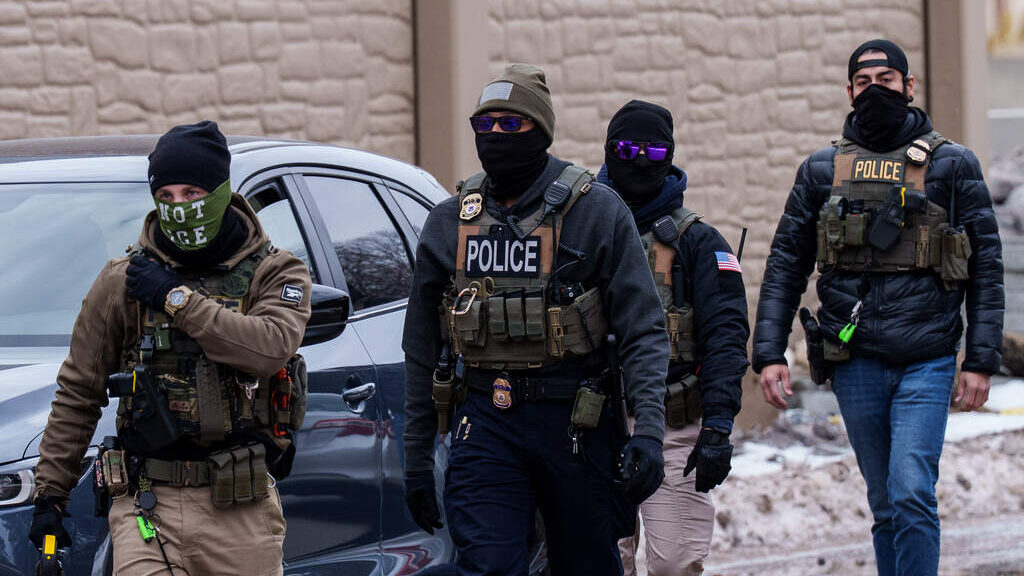 Four armed law enforcement officers in tactical gear and face coverings walking outdoors past a vehicle.